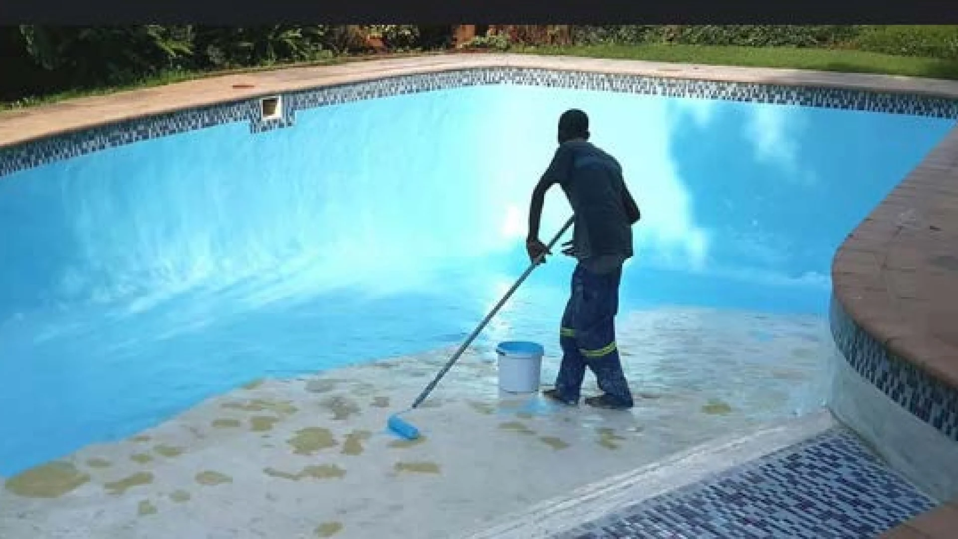 Worker applying epoxy paint inside an empty swimming pool.